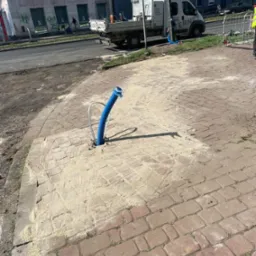 Niebieska rura wystająca z wywierconego otworu w chodniku z kostki brukowej, obsypana piaskiem, w tle ulica i budynki miejskie.