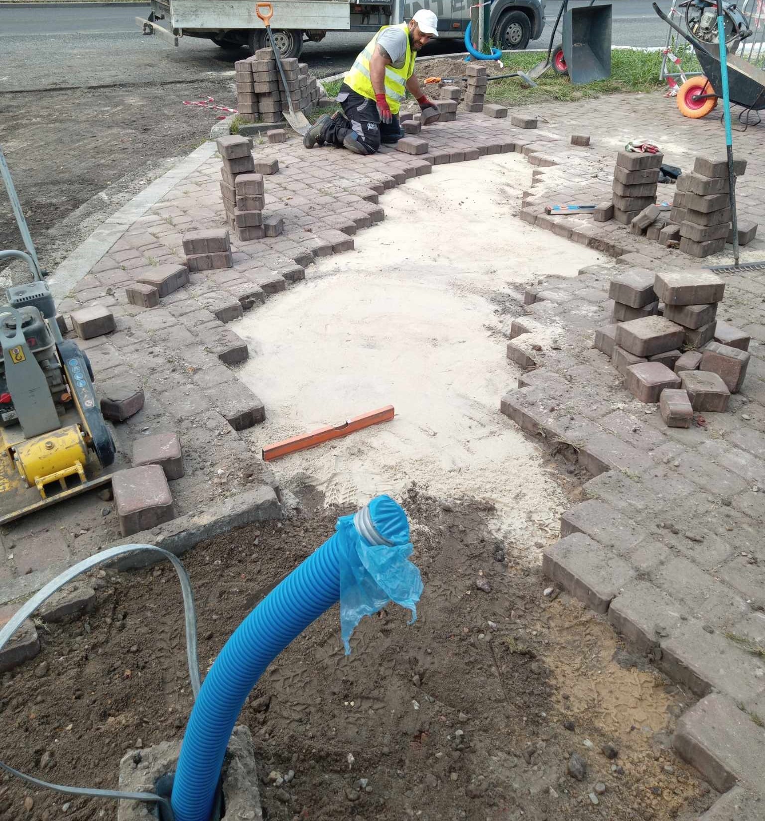 Prace brukarskie w toku: robotnik w kamizelce układa kostkę brukową na podsypce piaskowej. Widoczna zagęszczarka, stosy kostki, poziomica oraz niebieska rura instalacyjna wystająca z ziemi.