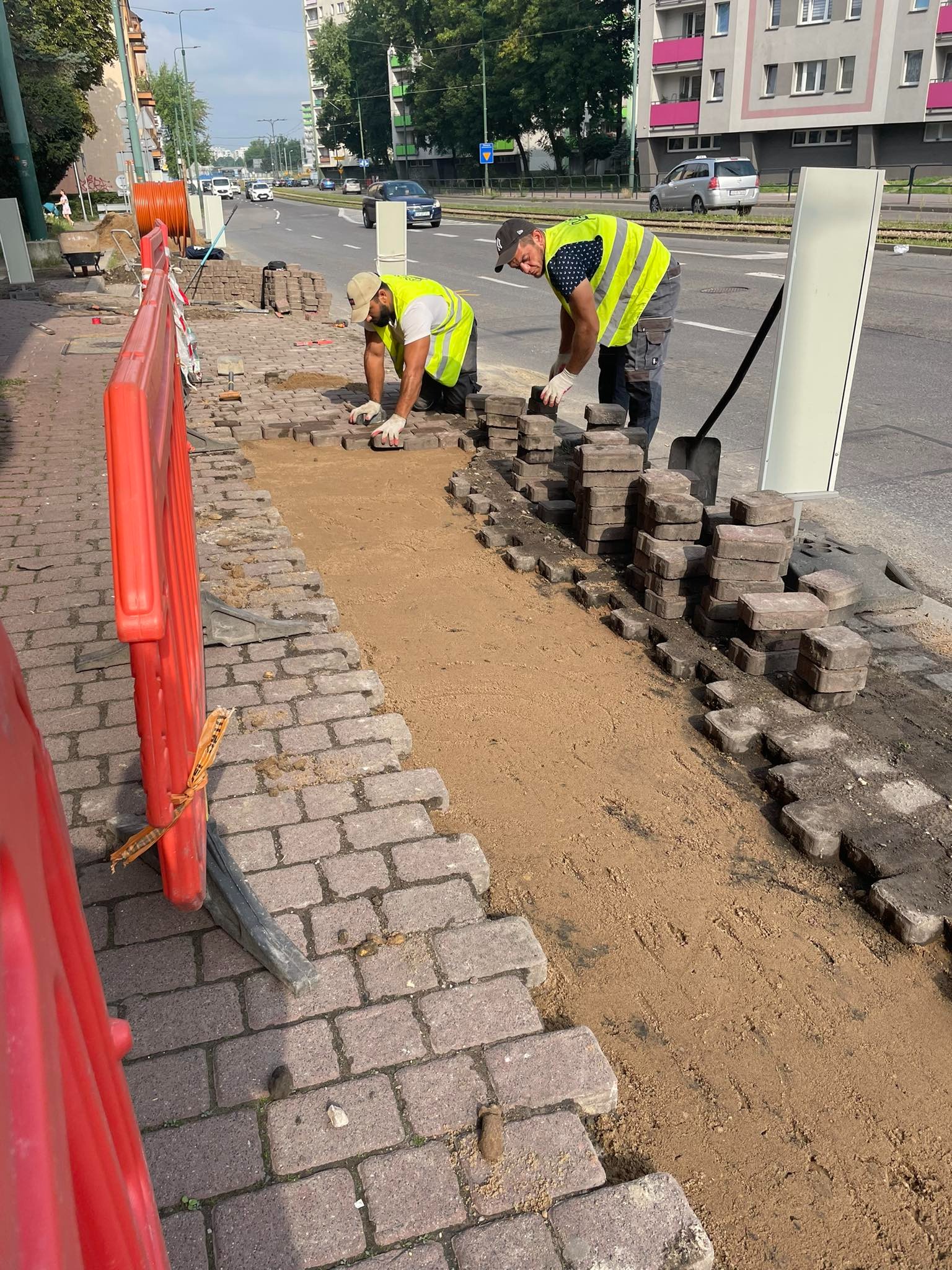 Dwóch robotników w kamizelkach odblaskowych układa kostkę brukową na chodniku przy ruchliwej ulicy miejskiej, widoczne porozrzucane kostki, piasek i czerwone bariery ochronne.