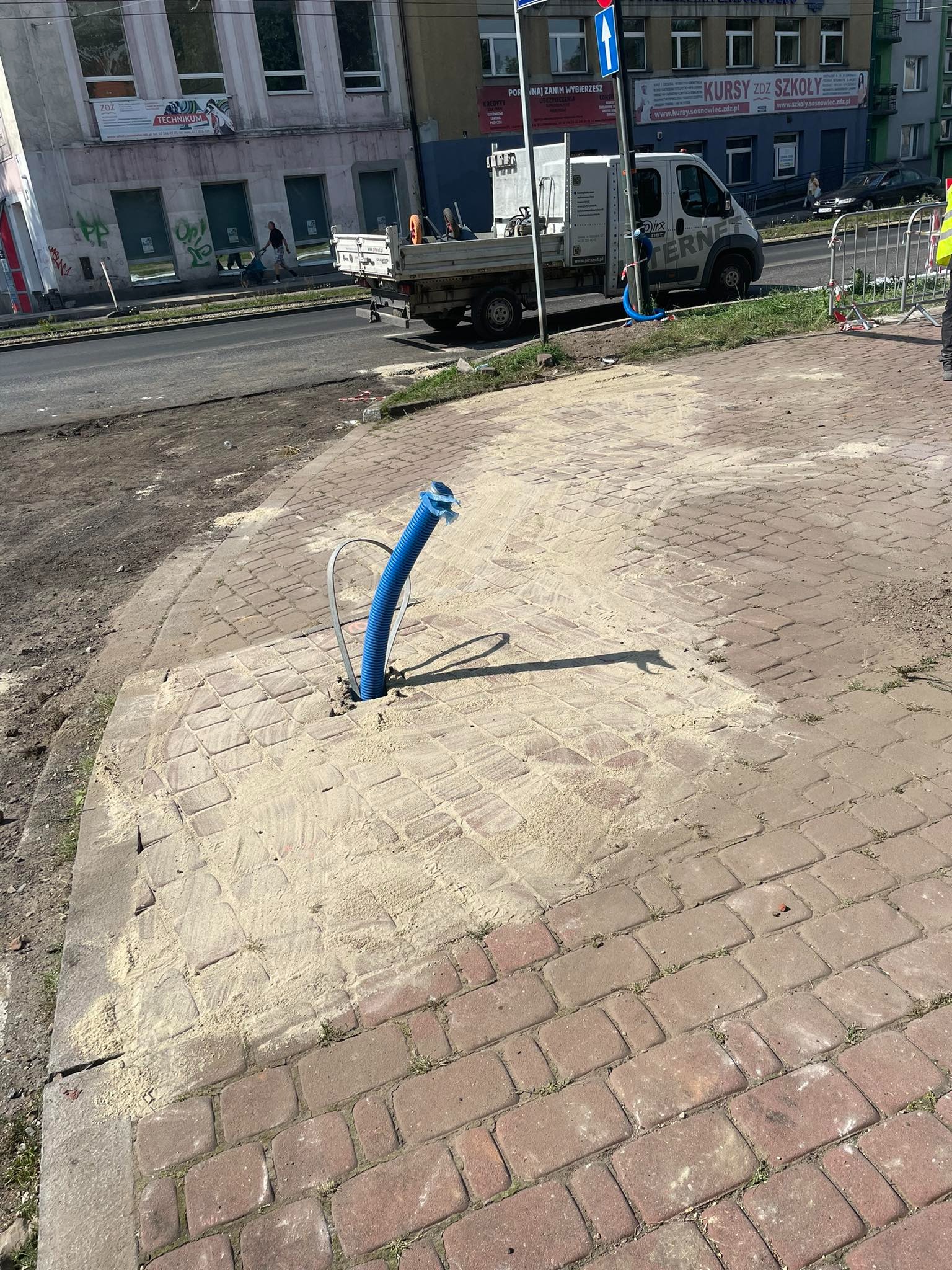 Niebieska rura wystająca z wywierconego otworu w chodniku z kostki brukowej, obsypana piaskiem, w tle ulica i budynki miejskie.