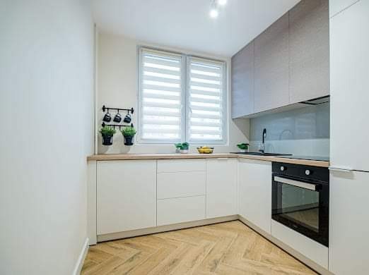 Jasna, minimalistyczna kuchnia z białymi frontami szafek, drewnianym blatem i podłogą w jodełkę; czarne akcenty w postaci piekarnika i relingów z doniczkami z ziołami.