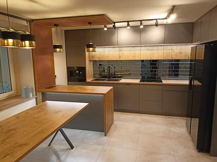 Nowoczesna kuchnia w odcieniach szarości z drewnianymi akcentami, wyspą i stołem z metalowymi nogami, oświetlona reflektorami i lampami wiszącymi.