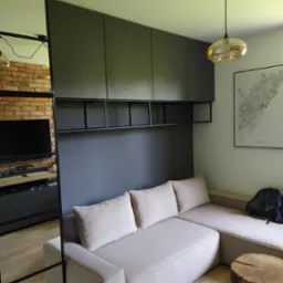 Beżowa sofa narożna z poduszkami, drewniany stolik-pień na metalowych nóżkach, zabudowa meblowa w kolorze grafitowym z otwartymi półkami, odbicie ściany z cegły w lustrze, mapa Barcelony na ścianie.