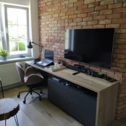Domowe biuro z blatem drewnianym na szafce RTV, ceglana ściana, laptop, fotel biurowy i stolik kawowy z pnia.