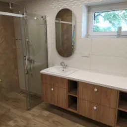 Nowoczesna łazienka z drewnopodobnymi płytkami podłogowymi i ściennymi, kabiną prysznicową ze szklanymi drzwiami przesuwnymi, szafką podumywalkową z drewnianymi frontami i białym blatem, owalnym...