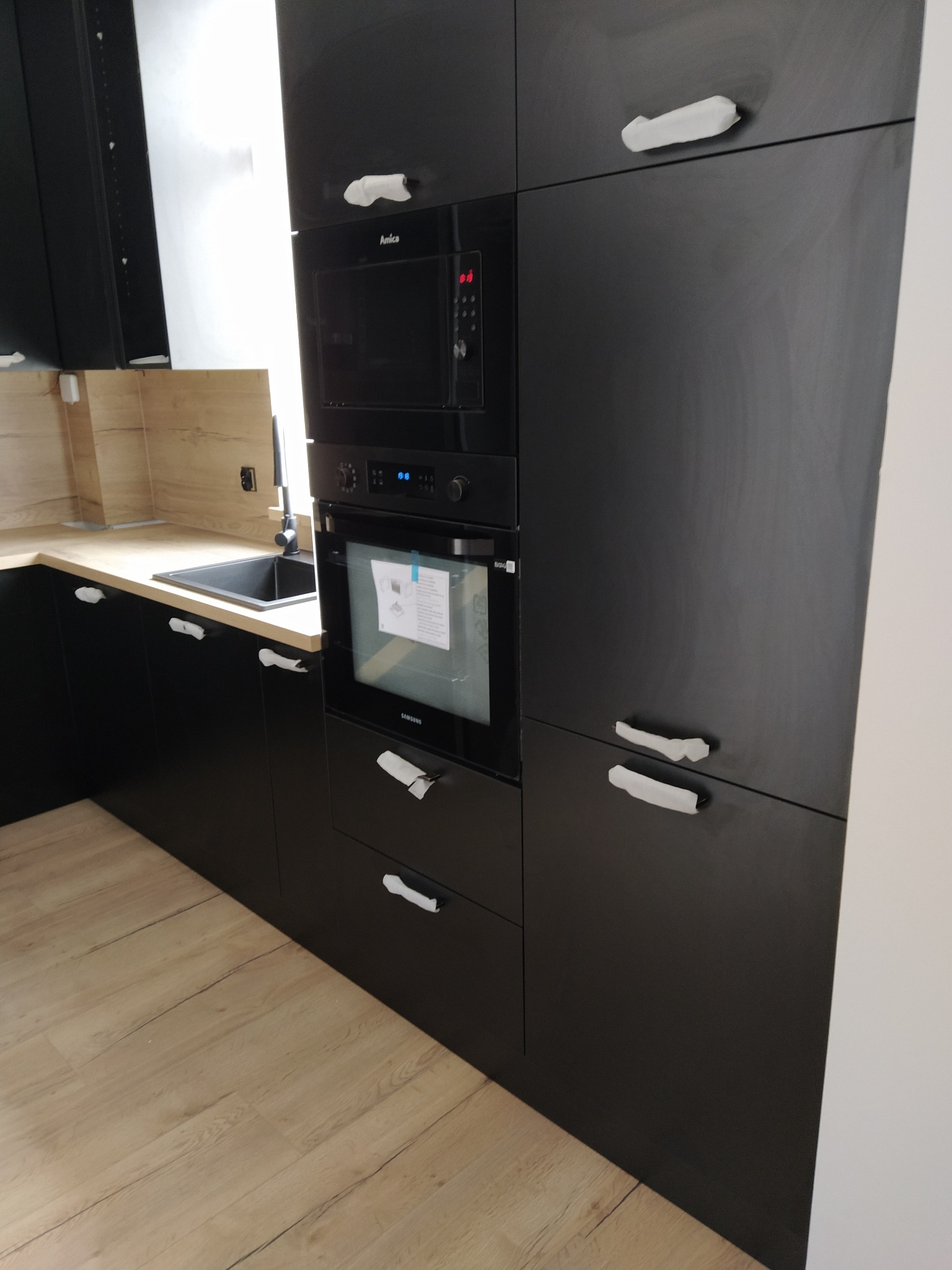 Częściowy widok nowej, czarnej zabudowy kuchennej z piekarnikiem i mikrofalówką w trakcie montażu, zabezpieczone uchwyty, jasna podłoga i fragment blatu roboczego.