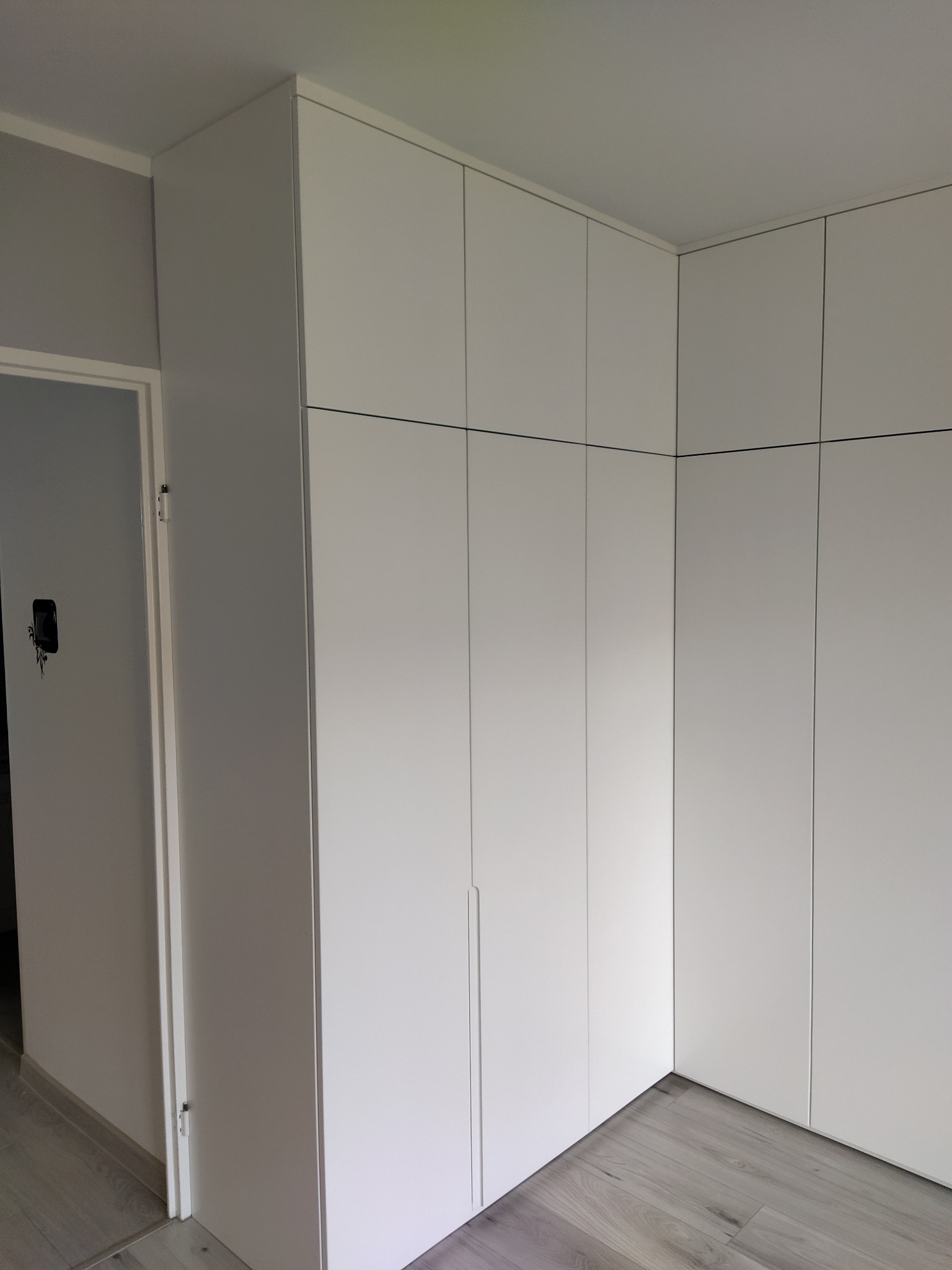 Biała, minimalistyczna szafa wnękowa zabudowana od podłogi do sufitu, z widocznymi pionowymi podziałami drzwi, na tle szarej ściany i podłogi z paneli imitujących drewno, widoczny fragment...