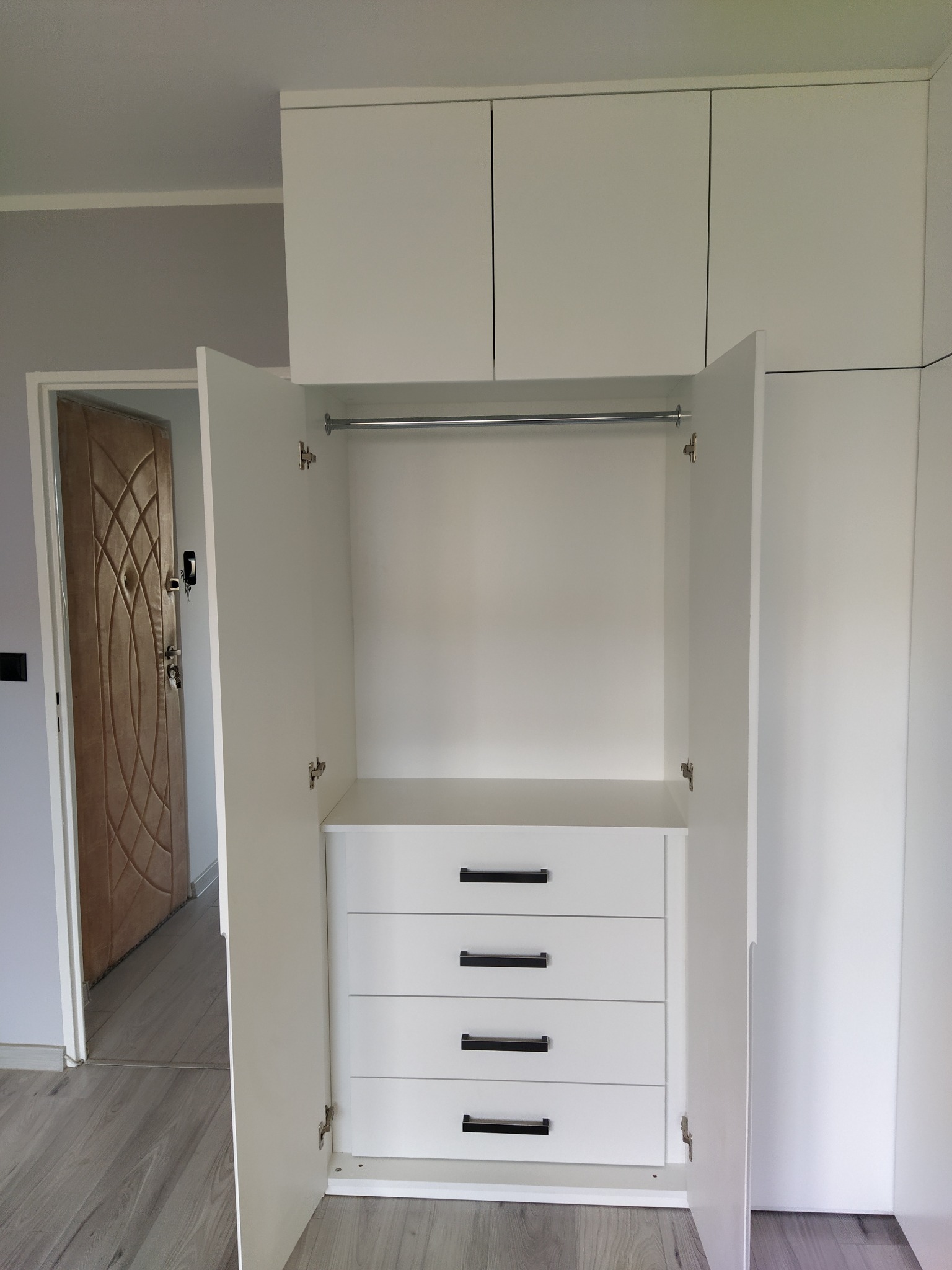 Wnętrze białej, otwartej szafy z czterema szufladami z czarnymi uchwytami i drążkiem na ubrania; widoczny fragment wejścia z brązowymi drzwiami.