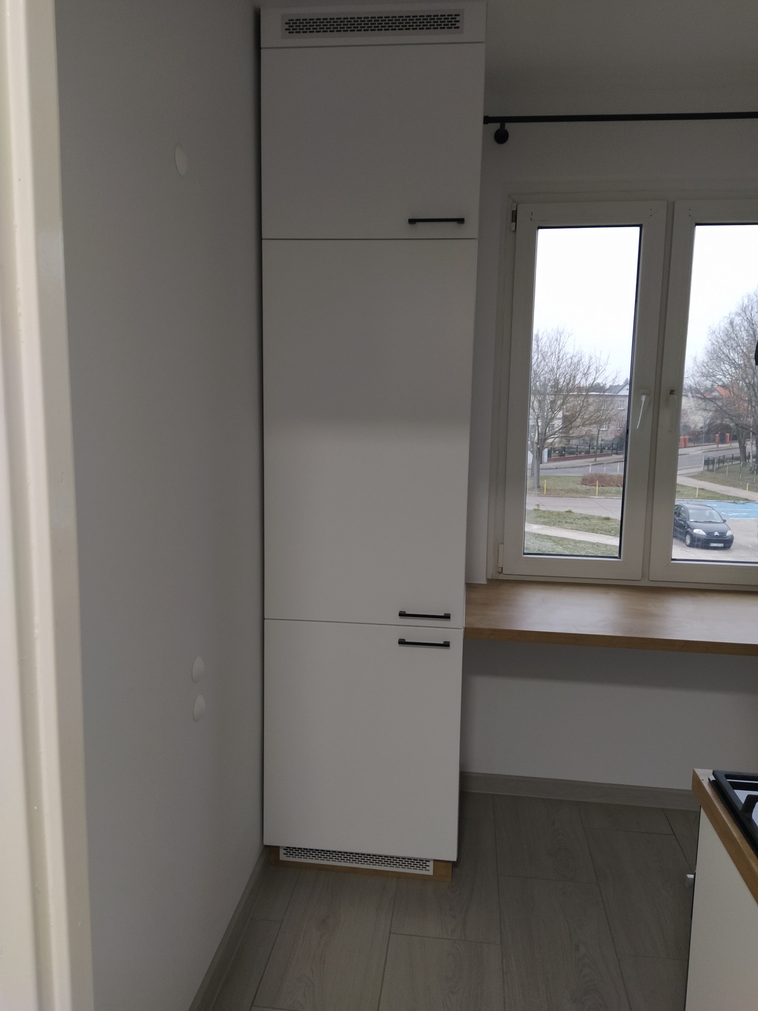 Wysoka, biała szafa w zabudowie kuchennej z czarnymi, minimalistycznymi uchwytami, umieszczona obok okna z drewnianym blatem, widok na osiedle z drzewami i zaparkowanym samochodem.