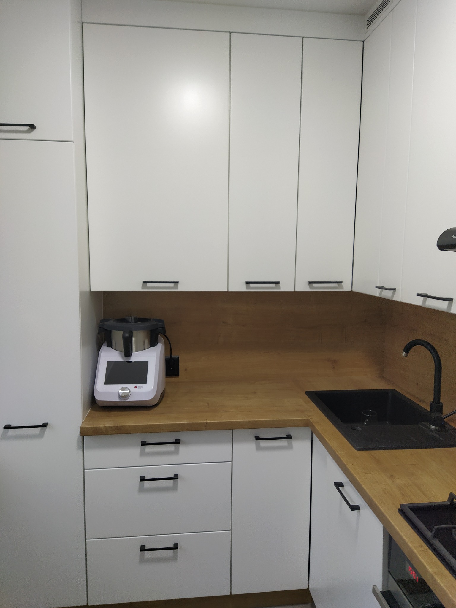 Nowoczesna, jasna kuchnia z białymi szafkami, drewnianym blatem i czarnym zlewem. Na blacie stoi biały robot kuchenny.