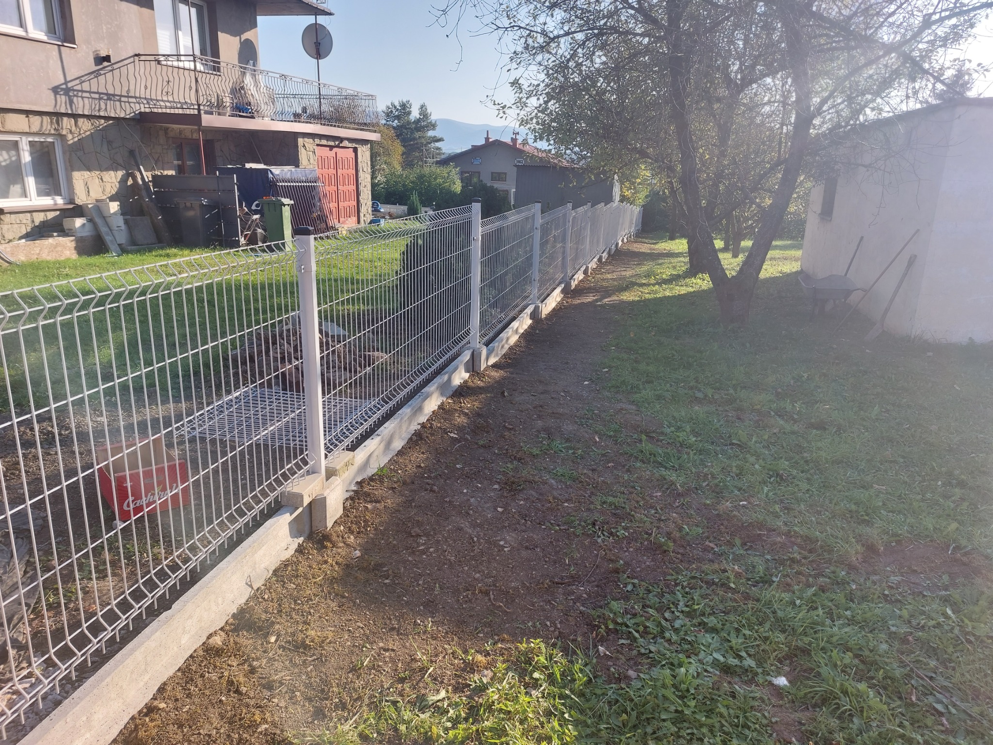 Długie, panelowe ogrodzenie z podmurówką betonową, widoczne z boku, ciągnące się wzdłuż działki z domem i ogrodem, z kartonem wewnątrz ogrodzenia.