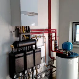Biały piec gazowy Vaillant zamontowany na ścianie, z widocznymi rurami miedzianymi i czerwonymi, podłączony do czarnego rozdzielacza z manometrami i białego zbiornika na wodę z niebieskim naczyniem...