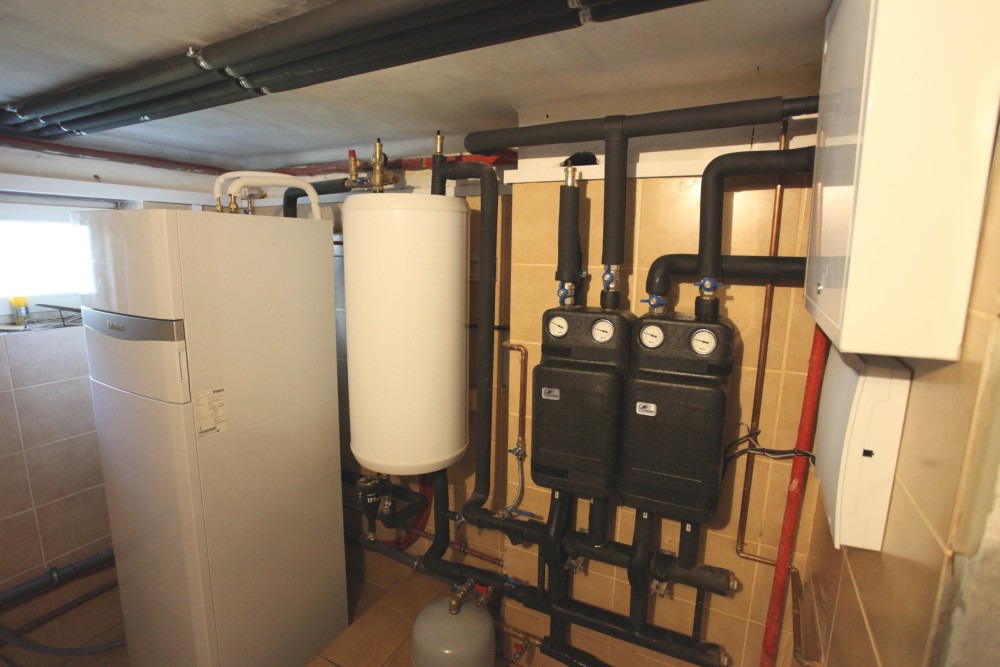 Biała pompa ciepła z zasobnikiem ciepłej wody i dwoma czarnymi modułami sterującymi na ścianie z beżowych płytek, widoczne rury instalacji grzewczej.