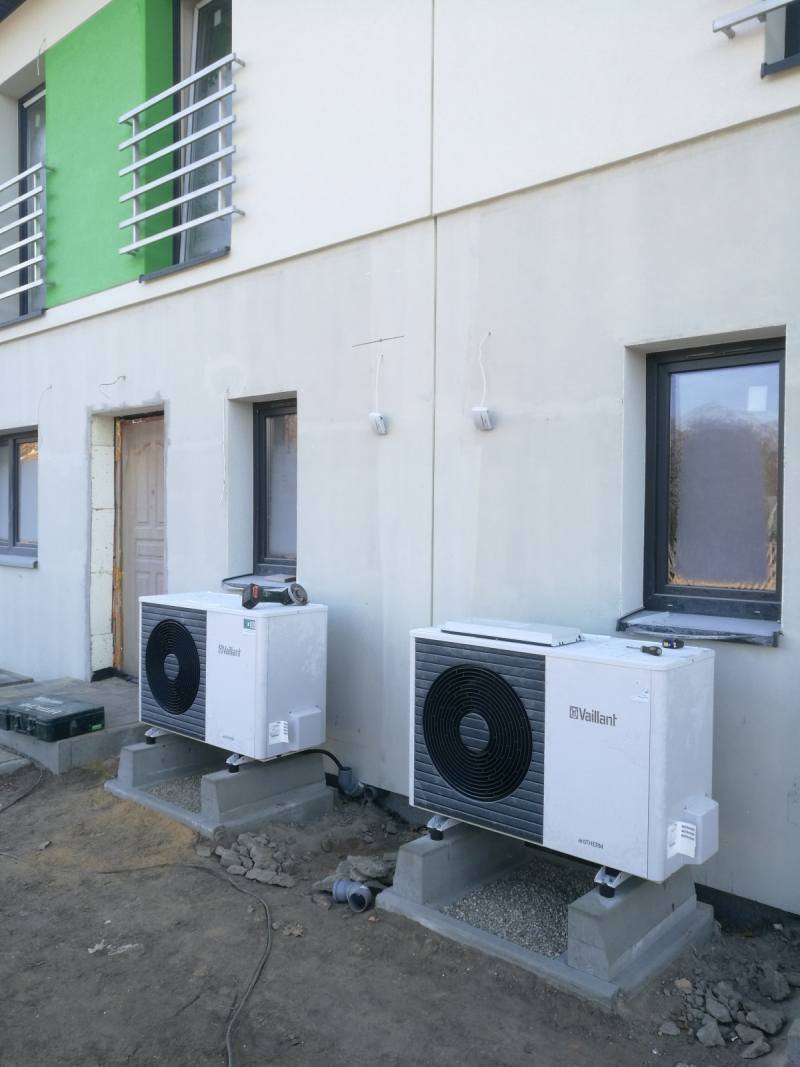 Dwie jednostki zewnętrzne marki Vaillant zamontowane na betonowych podstawach przy ścianie budynku z zielonym akcentem. Na jednej z jednostek leży szlifierka kątowa, a obok widoczna skrzynka...