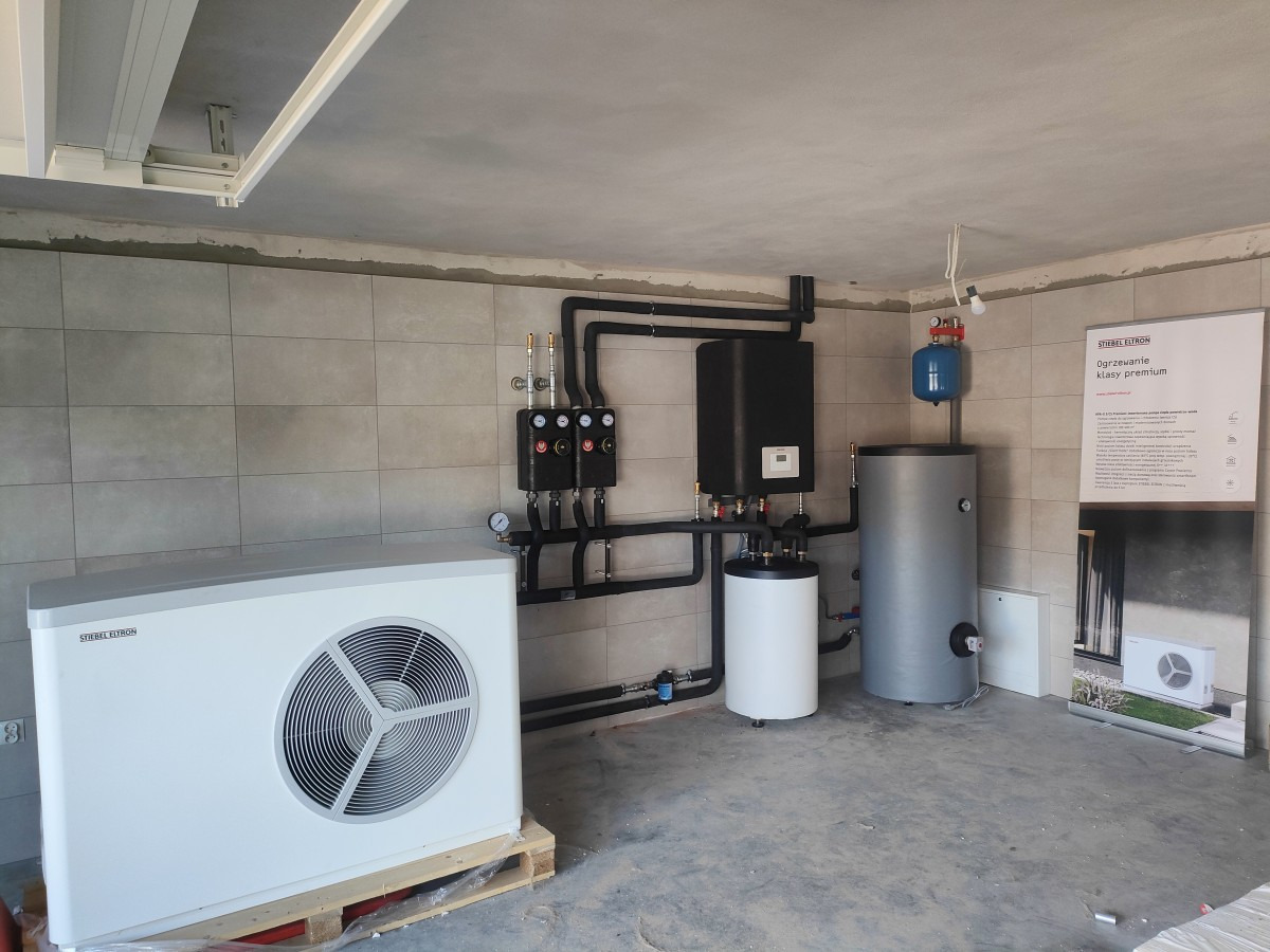 Zainstalowana pompa ciepła marki Stiebel Eltron na palecie w surowym wnętrzu, z widocznymi elementami instalacji grzewczej i zbiornikiem buforowym oraz plakatem informacyjnym.
