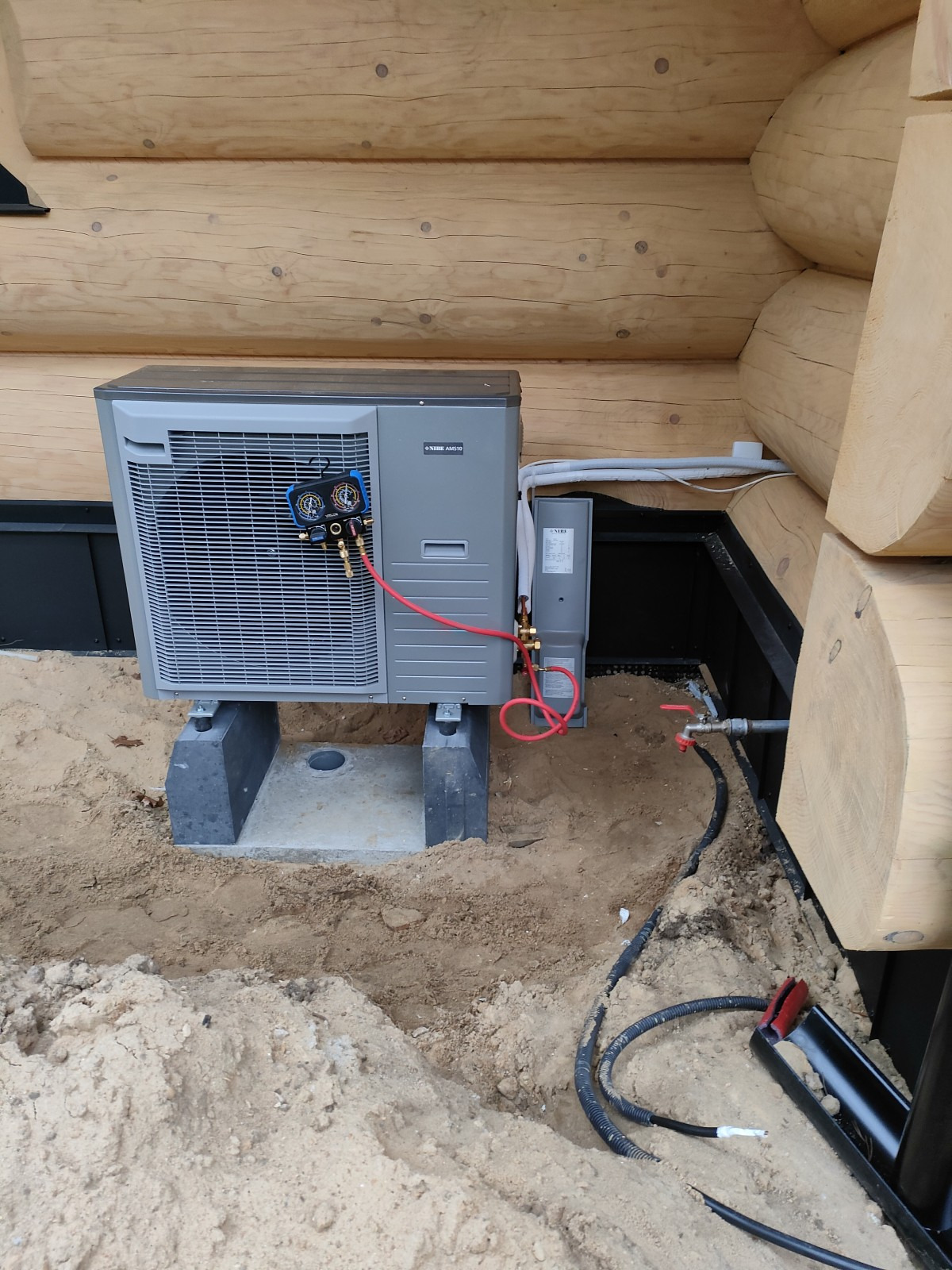 Szara jednostka zewnętrzna z podłączonymi przewodami pomiarowymi, umieszczona na betonowych wspornikach przy drewnianej ścianie domu z bali, widoczne rury i okablowanie w piasku.