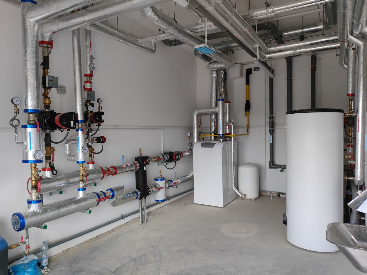 Kompletna kotłownia z piecem gazowym, zbiornikiem ciepłej wody użytkowej i rozbudowaną instalacją rur pokrytych izolacją, widoczne liczniki i zawory.