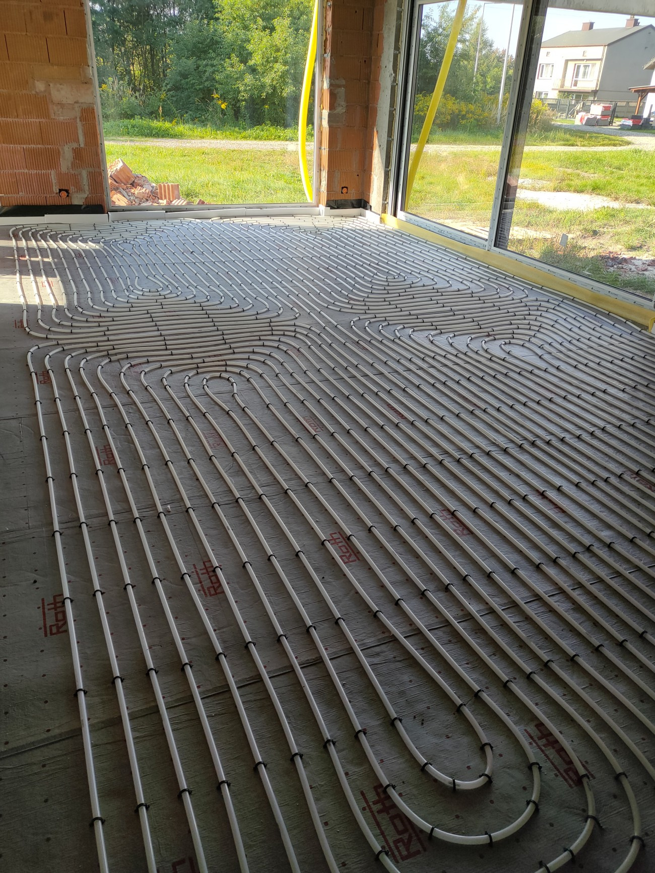 Rozłożone białe rury ogrzewania podłogowego z czarnymi łącznikami na szarej izolacji w surowym wnętrzu domu w trakcie budowy, widok z góry.