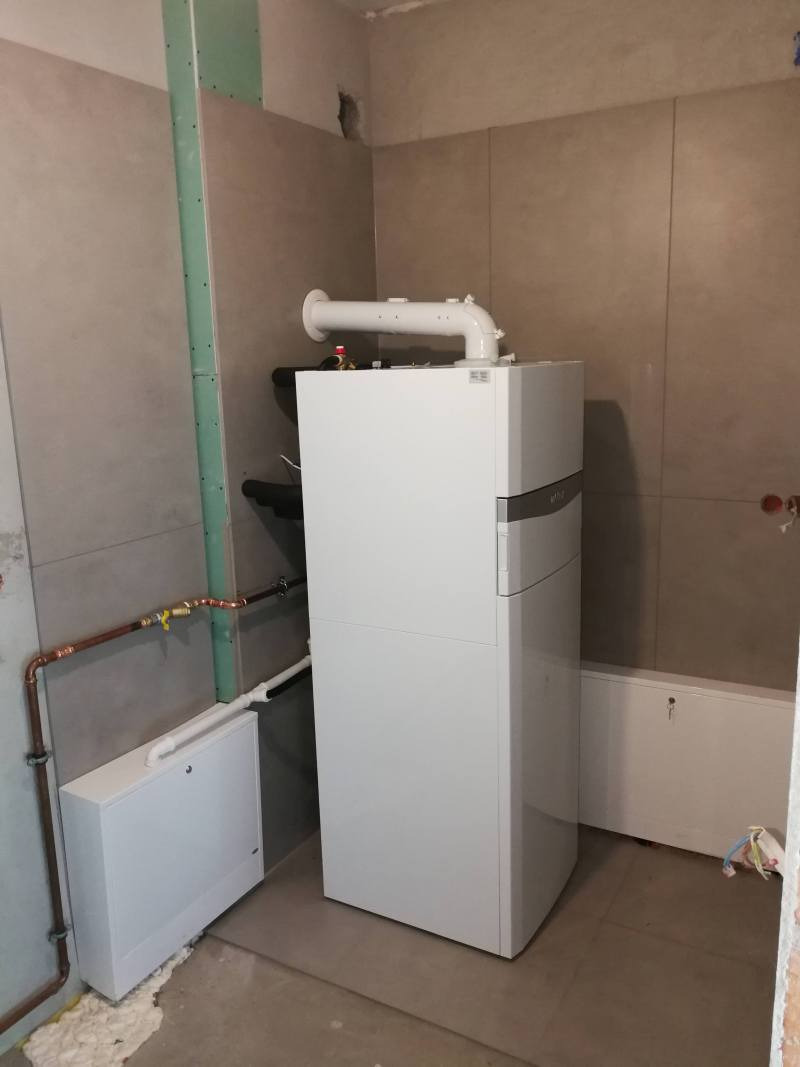 Biały piec gazowy kondensacyjny z podłączonymi rurami w trakcie instalacji w pomieszczeniu z szarymi płytkami i widocznymi śladami prac wykończeniowych.