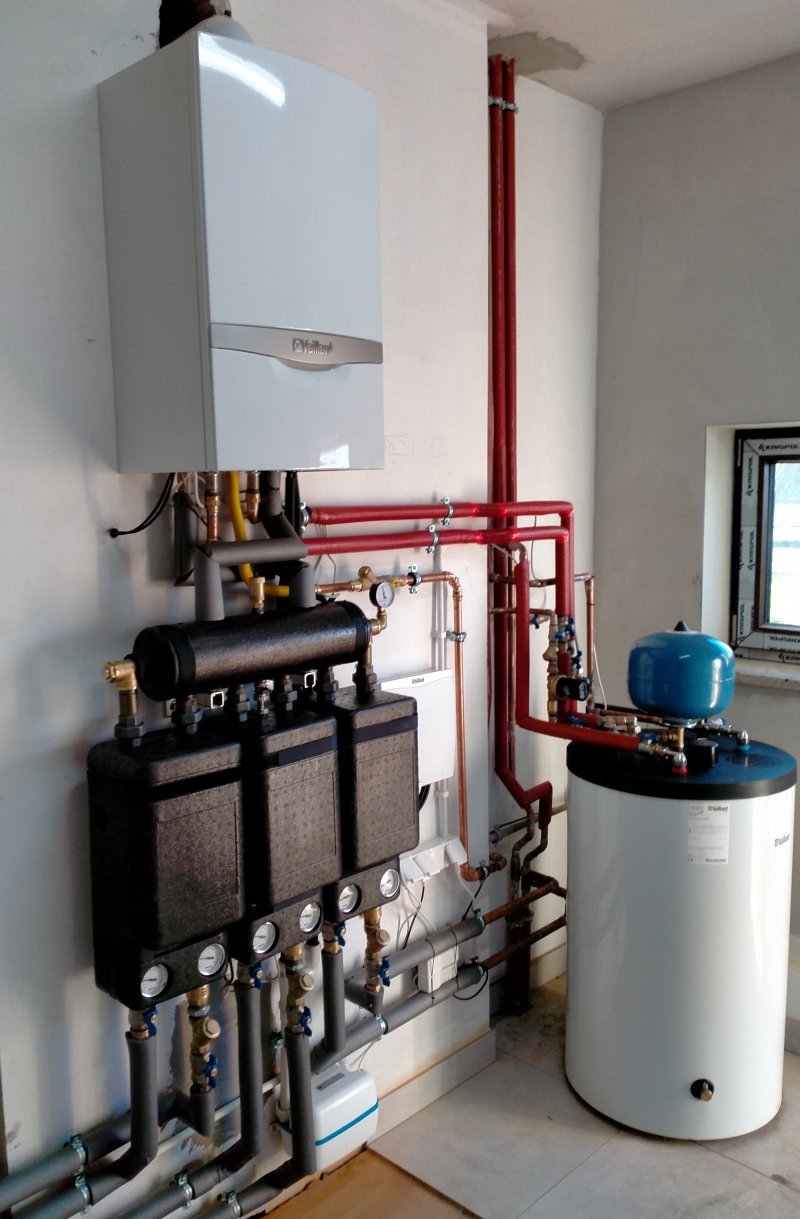 Biały piec gazowy Vaillant zamontowany na ścianie, z widocznymi rurami miedzianymi i czerwonymi, podłączony do czarnego rozdzielacza z manometrami i białego zbiornika na wodę z niebieskim naczyniem...