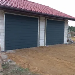 Dwa segmentowe, grafitowe wrota garażowe zamontowane w surowym budynku z białych bloczków, czerwony dach z blachodachówki, teren wokół wyrównany piaskiem, widoczna drabina oparta o ścianę.