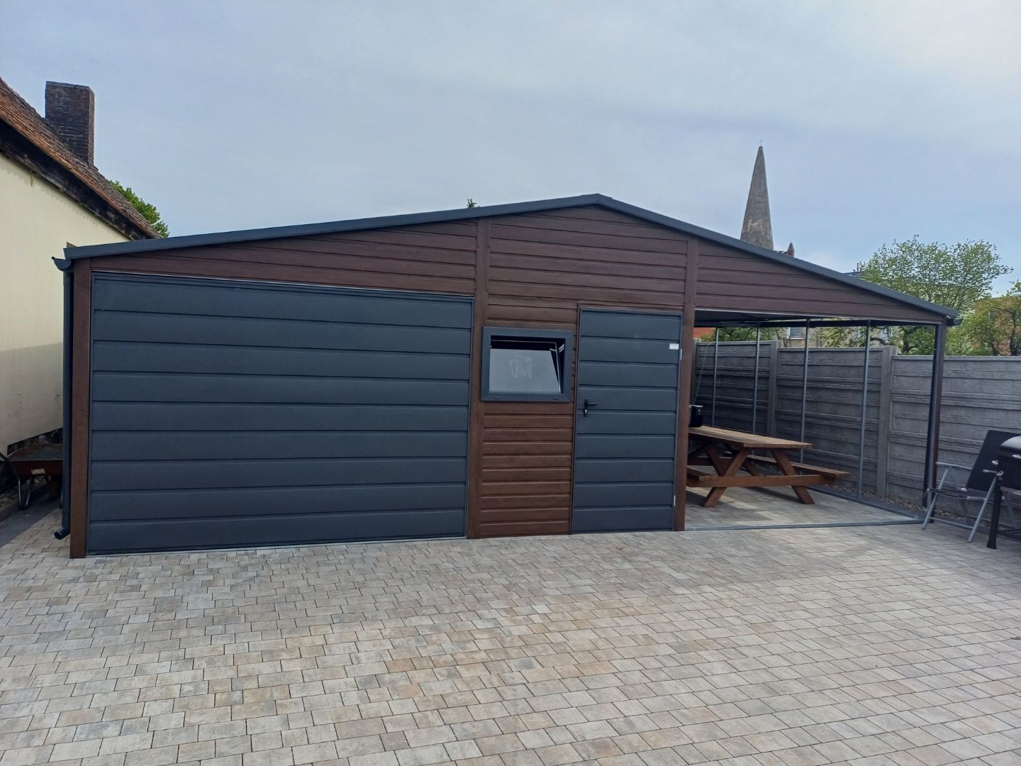 Nowoczesny garaż blaszany z bramą segmentową w kolorze antracytowym i częścią rekreacyjną z drewnianym stołem piknikowym, widok z zewnątrz na tle brukowanej nawierzchni.