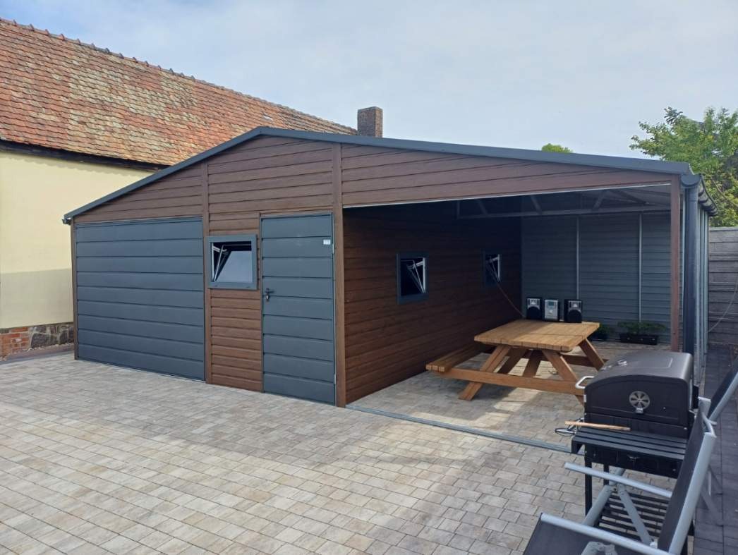 Dwukolorowy garaż z blachy trapezowej, część zamknięta z oknem i drzwiami, część otwarta z miejscem do grillowania i piknikowania, na kostce brukowej.