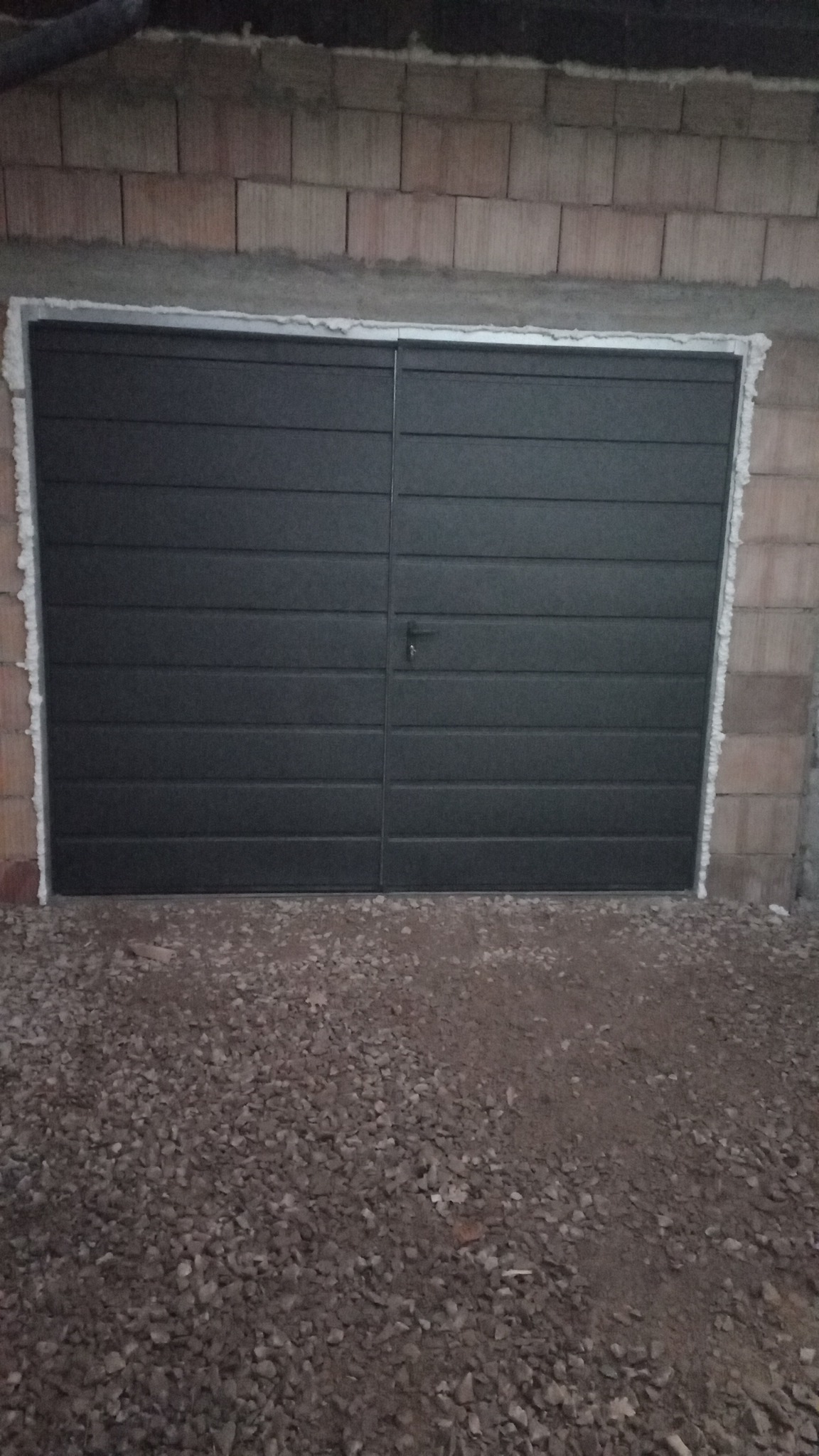 Zamknięta, dwuskrzydłowa, grafitowa brama garażowa z poziomymi przetłoczeniami, zamontowana w surowej ścianie z bloczków, widoczne uszczelnienie pianką montażową, podłoże z kamiennego grysu.