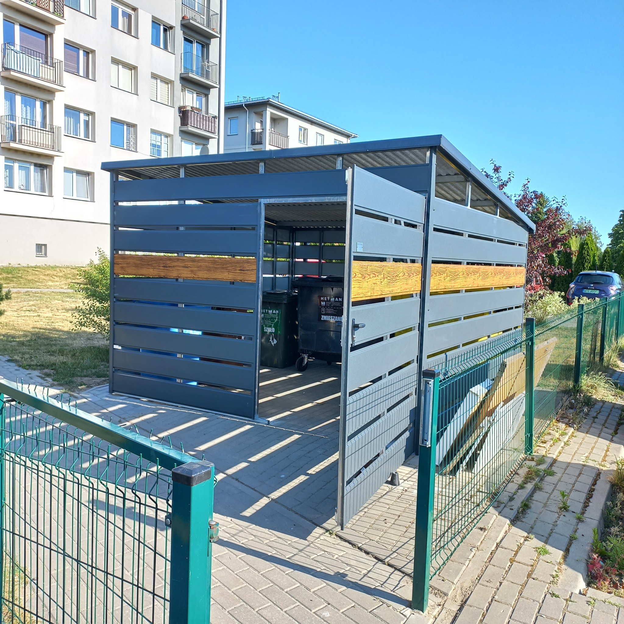 Szary, ażurowy schowek na pojemniki na odpady z drewnopodobnymi wstawkami, widoczne pojemniki na szkło i odpady zmieszane, na zewnątrz ogrodzenie z siatki.