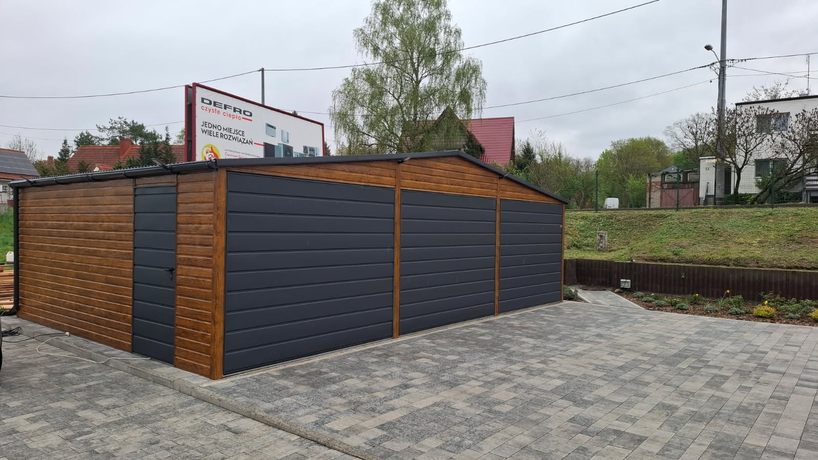 Gotowy, dwukolorowy garaż z blachy falistej, z bocznym wejściem i trzema bramami, na posesji wyłożonej kostką brukową, w tle reklama firmy Defro.