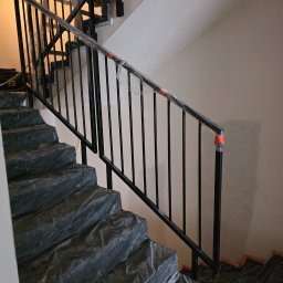 TECHNOMAR Marcin Kaczmarek - Nowoczesne, czarne schody metalowe z balustradą, zabezpieczone folią ochronną podczas montażu. Widok z dołu, jasne ściany w tle. Minimalistyczny design.