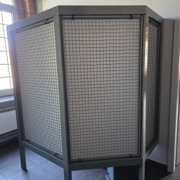 TECHNOMAR Marcin Kaczmarek - Metalowa konstrukcja z siatką, prawdopodobnie osłona lub obudowa, w szarym kolorze, ustawiona w pomieszczeniu z oknem i szarą podłogą. Widoczne elementy instalacji.