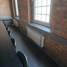 TECHNOMAR Marcin Kaczmarek - Grzejniki z metalową osłoną zamontowane na ceglanej ścianie pod oknami. Widoczne fragmenty czarnych krzeseł i stołu. Szara listwa przypodłogowa.
