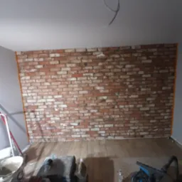 Ściana wykończona czerwoną cegłą dekoracyjną w trakcie prac, widoczne narzędzia i materiały budowlane, zabezpieczona podłoga drewniana.