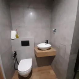 Szara łazienka z podwieszaną miską WC z bidetką, umywalką nablatową na drewnianym blacie i panelami podłogowymi o wzorze drewna.