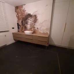 Nowoczesna łazienka z umywalką nablatową w kształcie łódki, szafką z poziomymi żłobieniami, marmurową ścianą w odcieniach brązu i beżu oraz czarną podłogą z żyłkami.