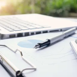 Laptop, pióro wieczne i kalkulator leżące na dokumencie z wykresami na jasnym biurku, w tle rozmyta zieleń.