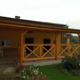 Zadaszony taras z drewnianą balustradą w kształcie krzyży, z widokiem na ogród z kwiatami, przy budynku z oknami i drzwiami.