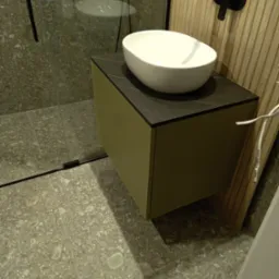 Nowoczesna umywalka nablatowa w kształcie misy, zamontowana na szafce w odcieniach oliwki z blatem imitującym ciemny marmur, czarna bateria ścienna, kabina prysznicowa z szarymi płytkami w tle.