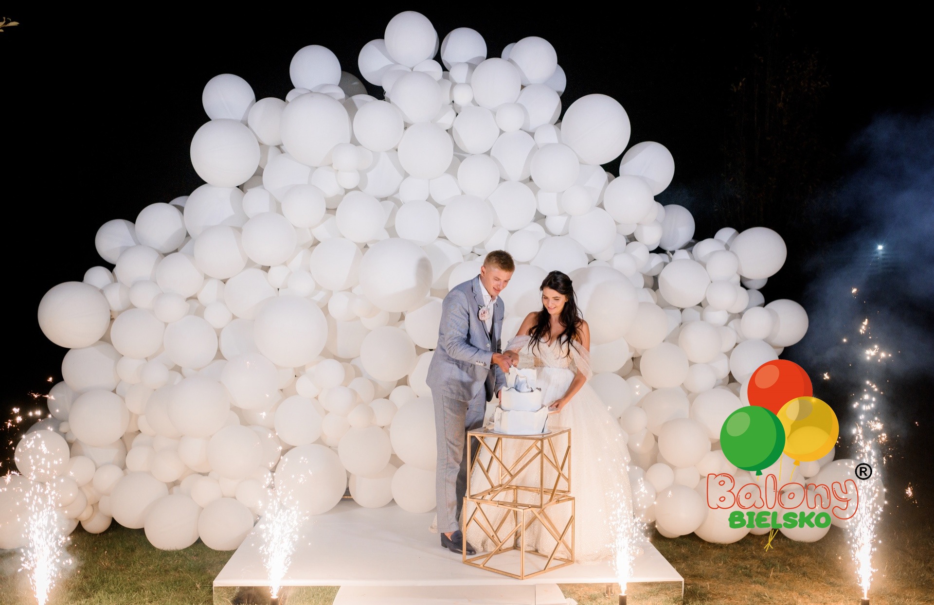 Młoda para kroi tort weselny na tle ściany z białych balonów i fontanny iskier.