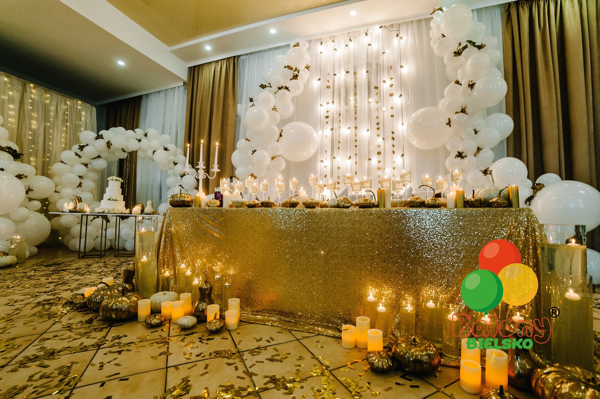Uroczysta dekoracja stołu z błyszczącym złotym obrusem, białymi balonami, świecami, dyniami i złotymi konfetti na podłodze.