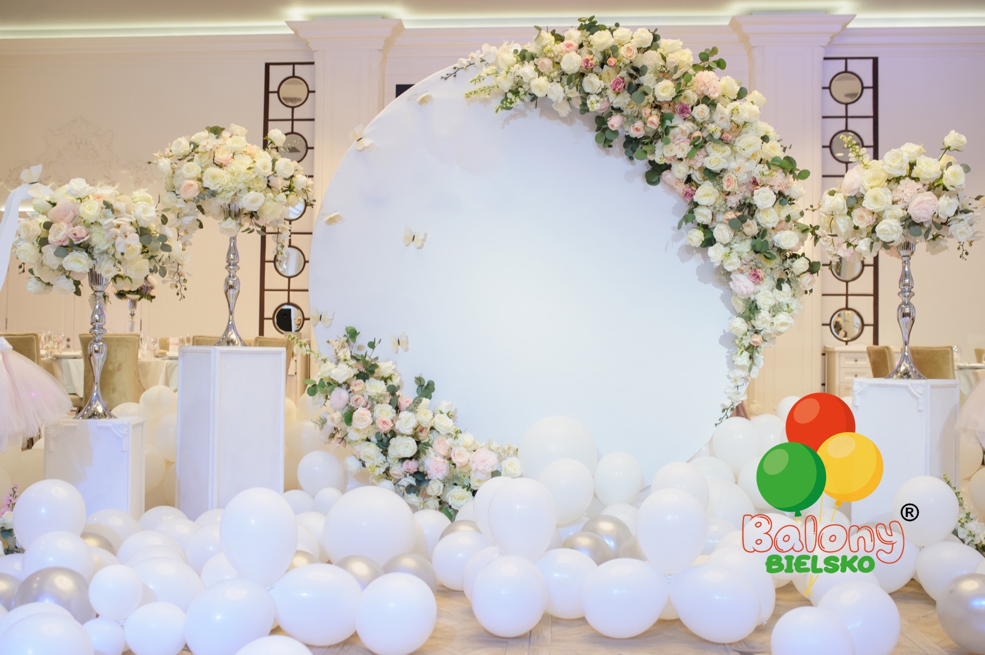 Dekoracja sali bankietowej: białe balony na podłodze, okrągła ścianka z kwiatami, wysokie wazony z kompozycjami kwiatowymi, logo firmy z balonami w prawym dolnym rogu.