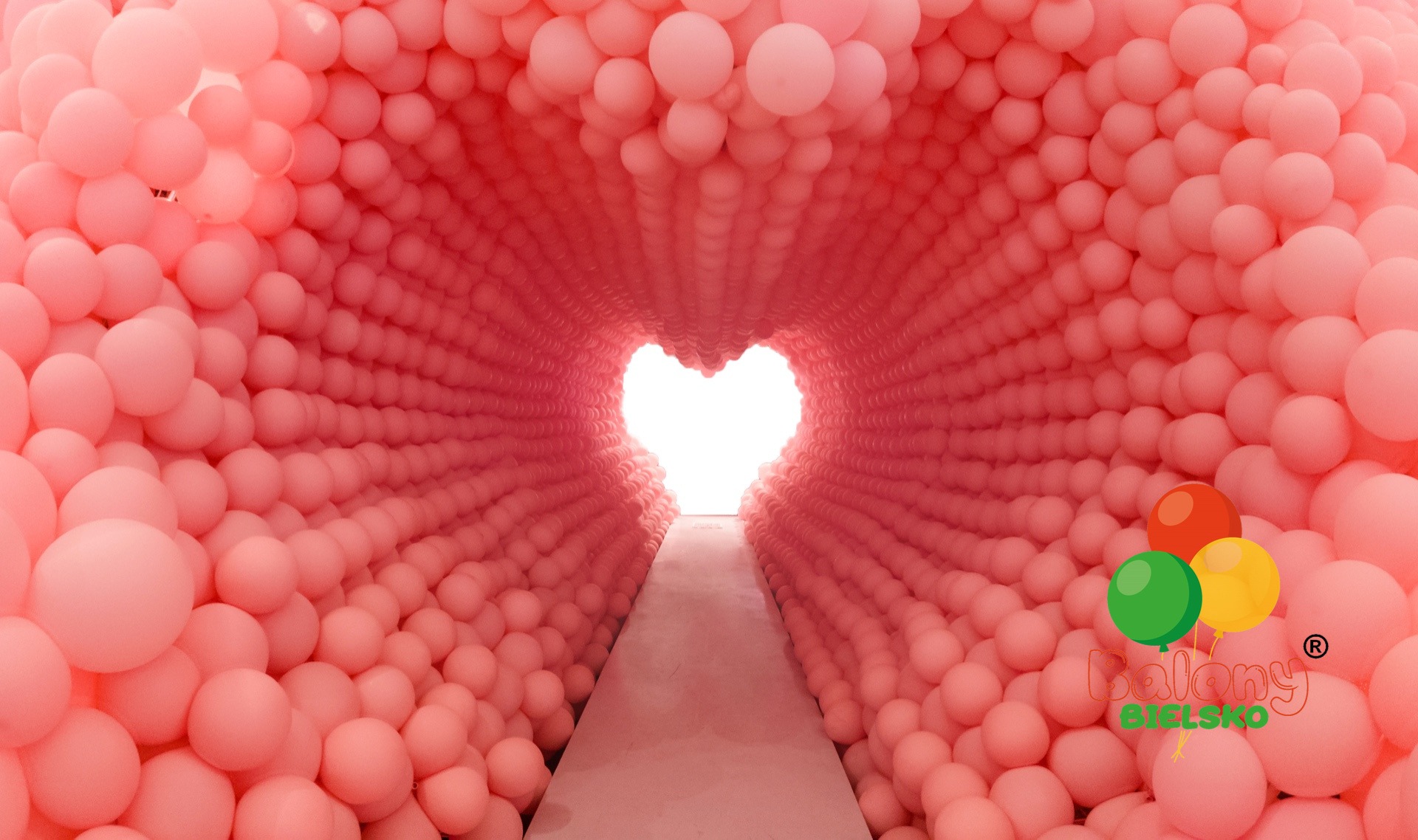 Tunel z różowych balonów prowadzący do jasnego, sercowatego otworu, z logo firmy balonowej w prawym dolnym rogu.