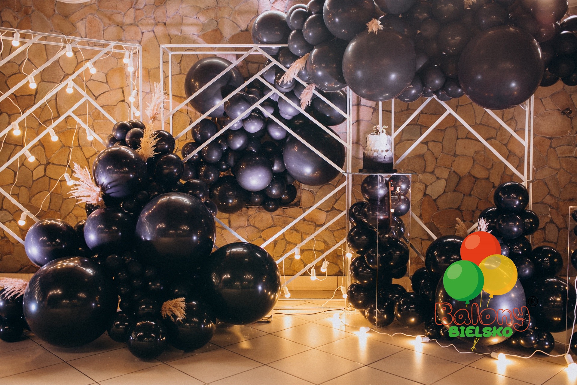 Dekoracja sali czarnymi balonami z elementami suchej trawy pampasowej, podświetlona girlandą żarówek, z tortem na przezroczystym postumencie w tle.