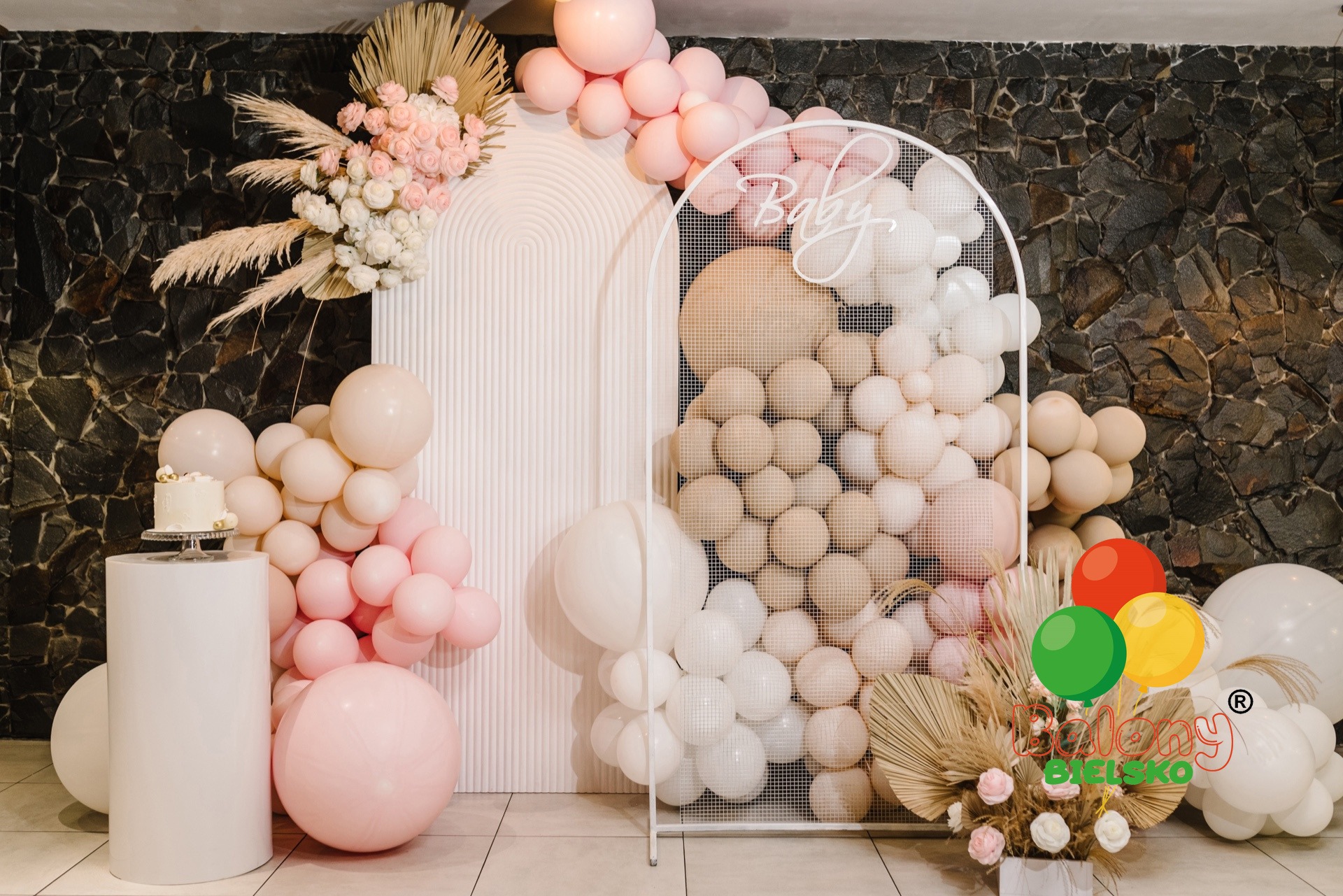 Dekoracja balonowa w odcieniach pastelowych różów i beżów z napisem 'Baby' na tle ściany z kamienia, tort na białym postumencie, kompozycja z suszonych traw i kwiatów.