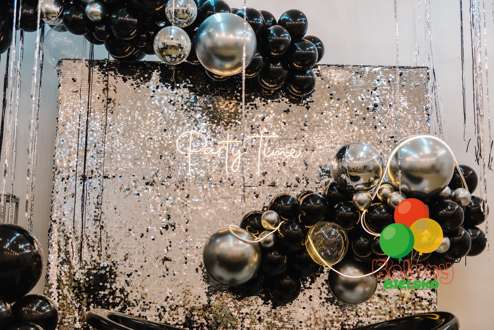 Dekoracja imprezowa: błyszcząca ścianka z cekinami, neonowy napis 'Party Time' oraz girlanda z czarnych, srebrnych i przezroczystych balonów z dodatkiem kolorowych balonów w dolnej części kompozycji.