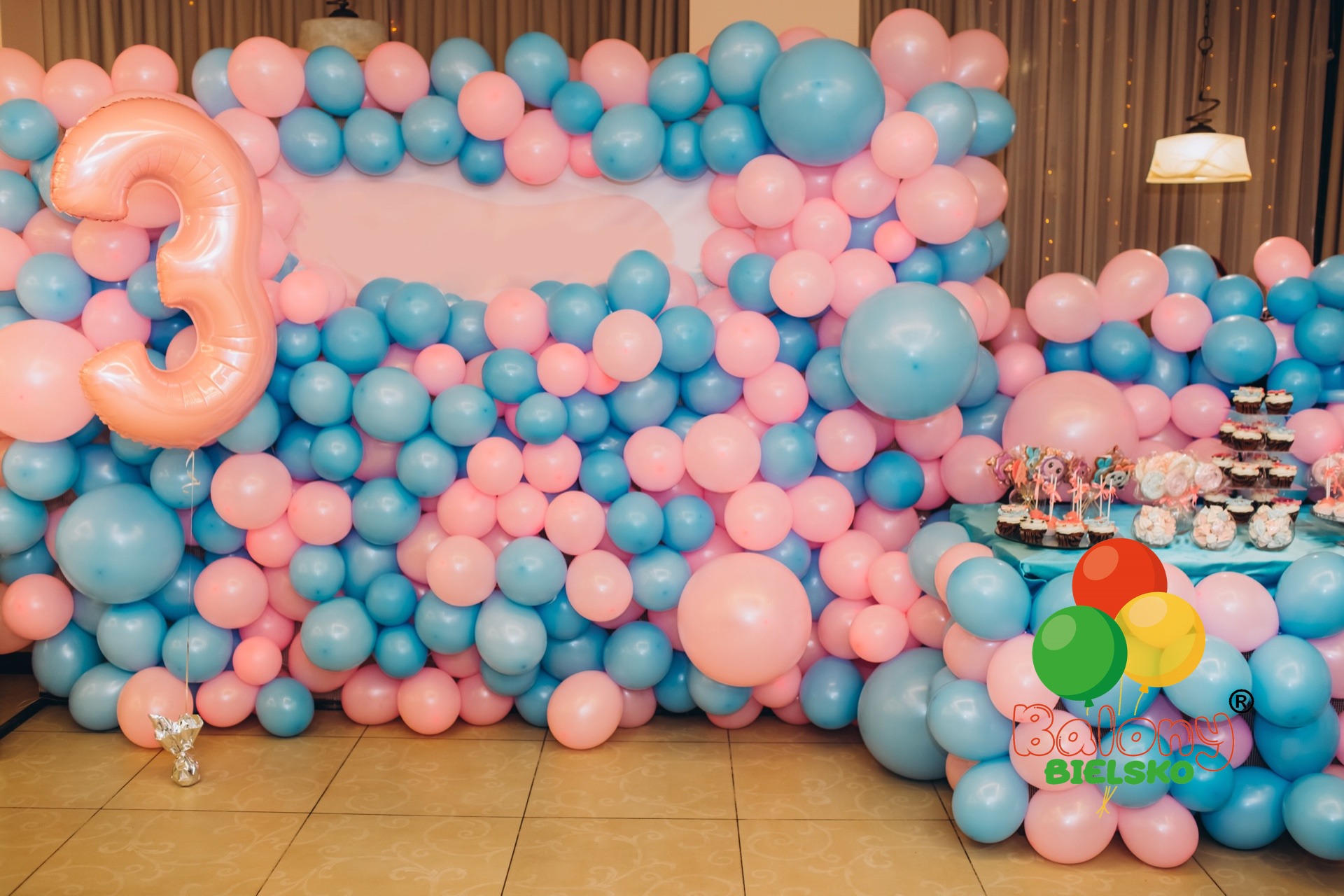 Dekoracja z niebieskich i różowych balonów z cyfrą '3' z folii, słodki stół z babeczkami i lizakami w tle, logo firmy balonowej w prawym dolnym rogu.