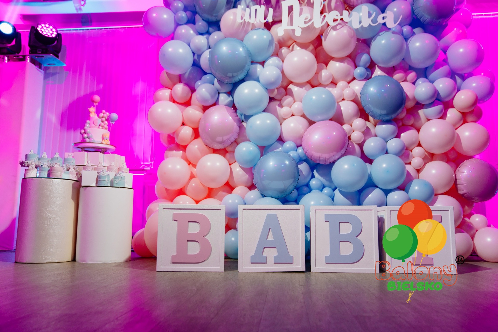 Dekoracja urodzinowa z balonów w odcieniach różu i błękitu, tort i babeczki na podestach, drewniane litery układające się w słowo 'BABY' oraz kolorowe balony.