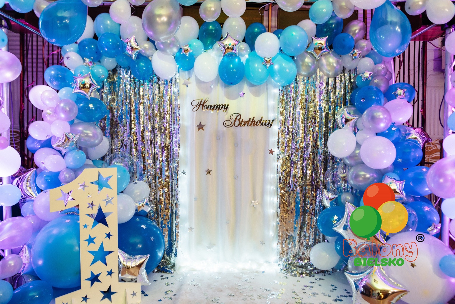 Dekoracja na pierwsze urodziny z niebiesko-białymi balonami, srebrnymi gwiazdami i błyszczącą kurtyną, z napisem 'Happy Birthday' i liczbą '1' z wyciętymi gwiazdami.