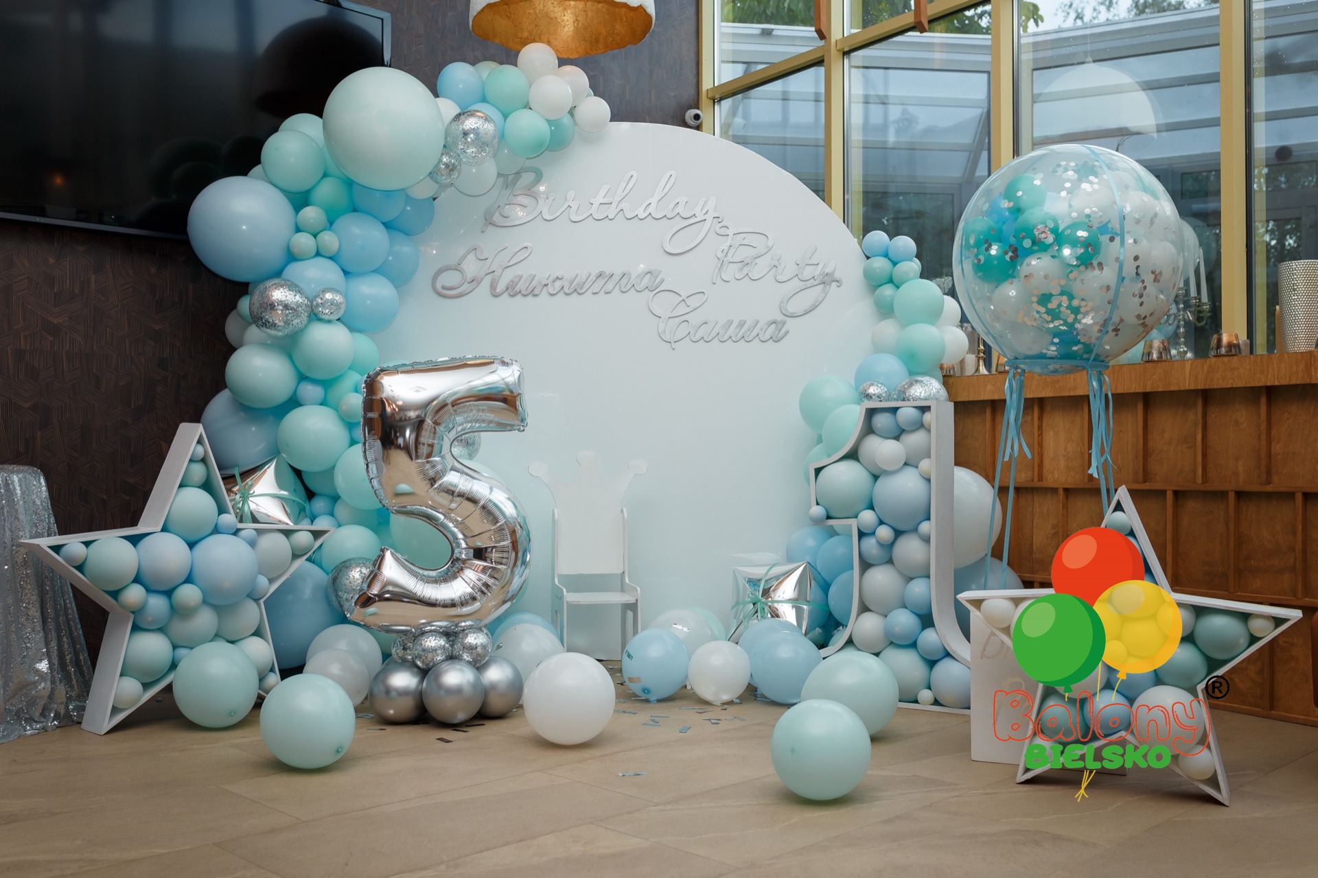 Dekoracja balonowa w odcieniach błękitu i srebra na piąte urodziny, z napisem 'Birthday Huruma Party Gama' na białym tle, gwiazdami wypełnionymi balonami i balonem foliowym w kształcie cyfry 5.